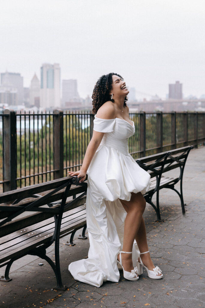 Bride in an elegant off-shoulder dress, laughing joyfully while leaning against a bench at the Brooklyn Heights Promenade with the NYC skyline in the background