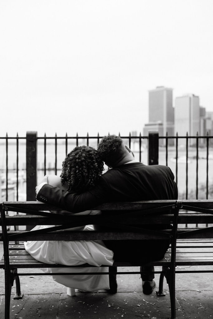 Black-and-white photo of a bride and groom sitting on a bench, sharing a quiet moment while overlooking the NYC skyline during their Brooklyn elopement