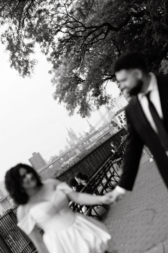 Black-and-white motion blur photo of a bride and groom holding hands, walking at Brooklyn Heights Promenade with the NYC skyline in the background.