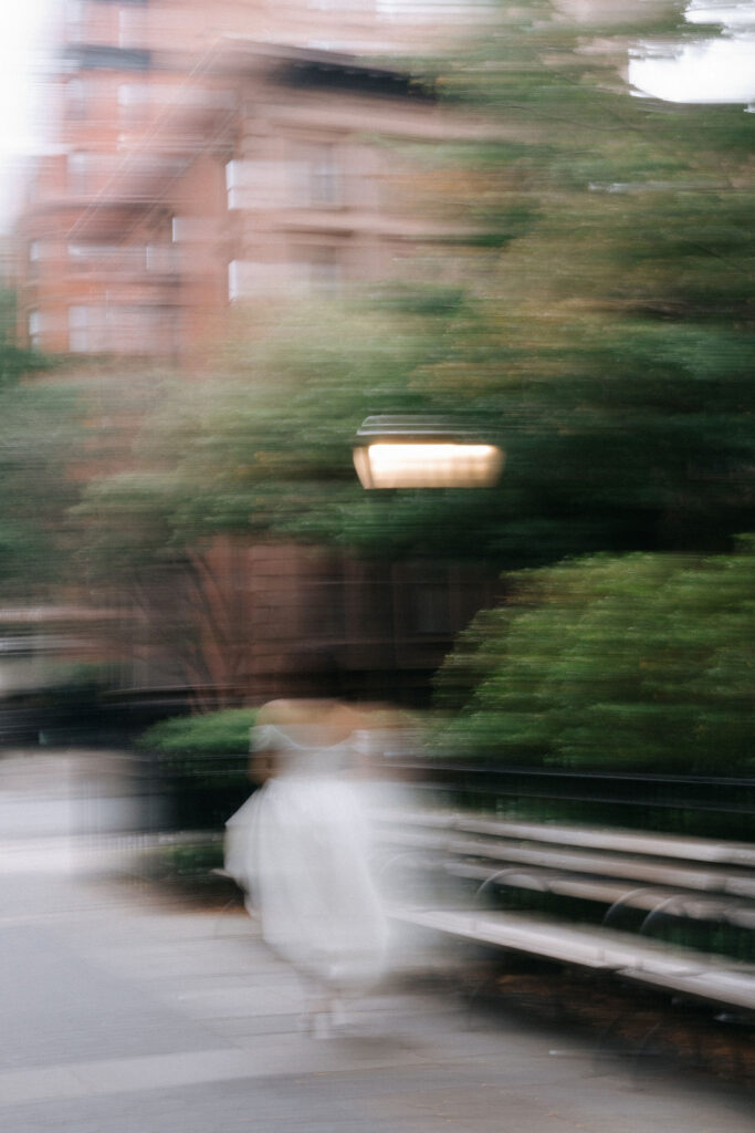 Artistic motion blur of a bride in an off-the-shoulder wedding dress walking through a quiet Brooklyn street, surrounded by soft greenery and urban charm.