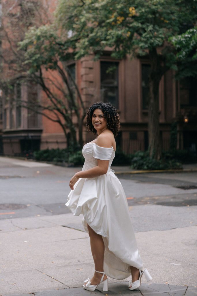 Elegant bride in an off-the-shoulder gown smiling on a quiet, tree-lined Brooklyn street, surrounded by brownstone architecture.