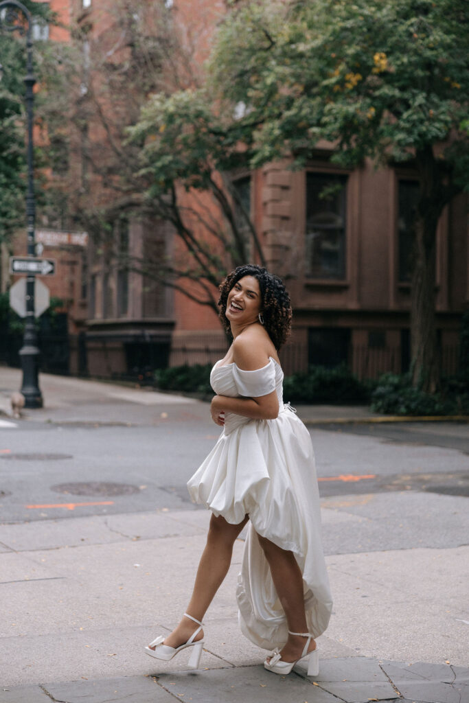 Happy bride in an off-the-shoulder gown smiling and walking down a quiet, picturesque Brooklyn street, surrounded by classic brownstones and greenery