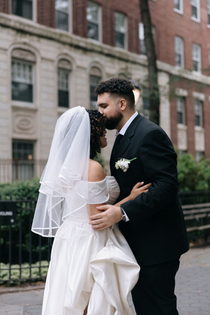 Groom gently kisses the bride's forehead during an intimate moment in a charming Brooklyn neighborhood, with classic urban architecture in the background.