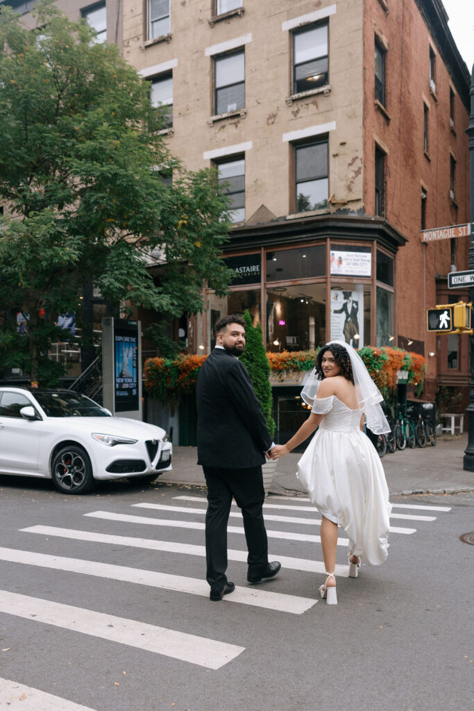 Bride and groom holding hands and smiling as they cross a street in Brooklyn, surrounded by charming urban architecture and a cozy café.