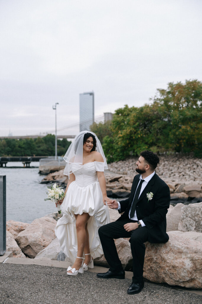 A bride and groom sharing a moment by the waterfront in Brooklyn, with the bride standing gracefully on rocks and the groom seated, looking up at her with admiration