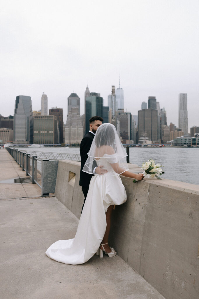 A bride and groom overlooking the NYC skyline from Brooklyn, with the bride leaning on a concrete barrier holding her bouquet and the groom standing close behind her.