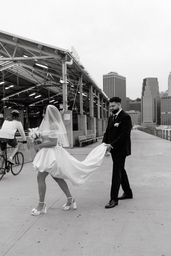 Black and white image of a groom gently holding the train of his bride's dress as they walk along Brooklyn's waterfront pier, with NYC buildings in the background