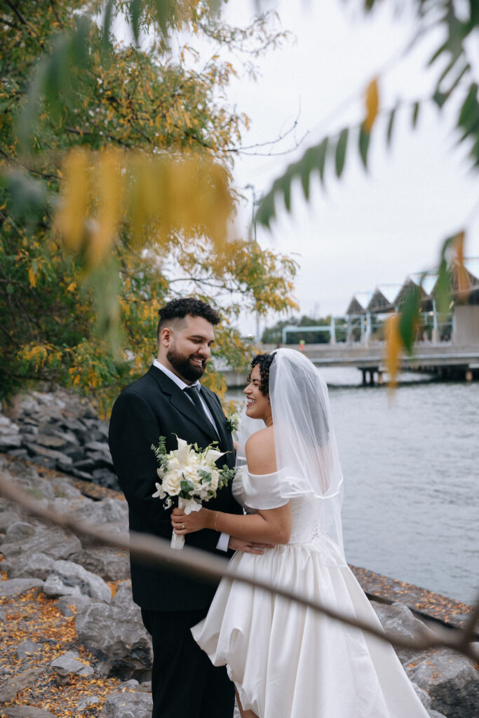 Romantic moment of a bride and groom standing by the waterfront, surrounded by autumn leaves and soft greenery, sharing a joyful glance