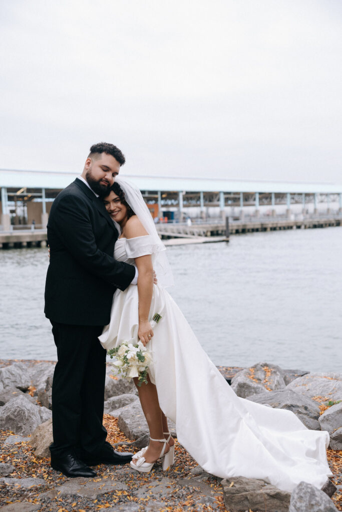 Newlywed couple embracing on a rocky waterfront, the bride holding a bouquet and wearing an off-the-shoulder gown with a long train.