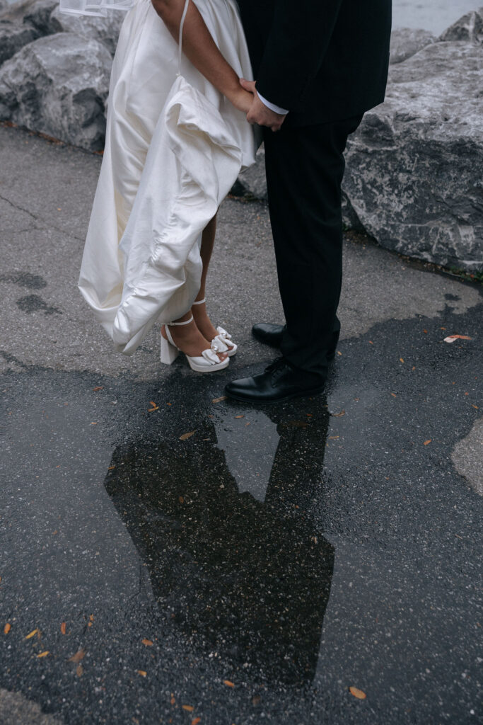 Close-up of a bride and groom holding hands, their reflection captured in a puddle on a rocky waterfron