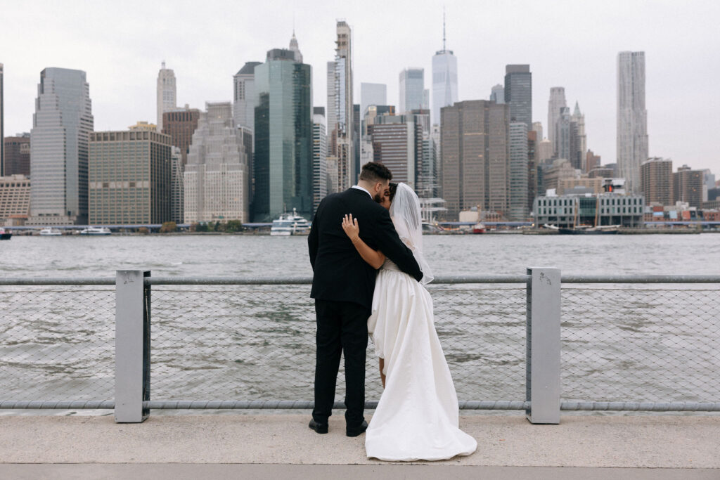 Couple embracing while standing by the waterfront with the Manhattan skyline in the background