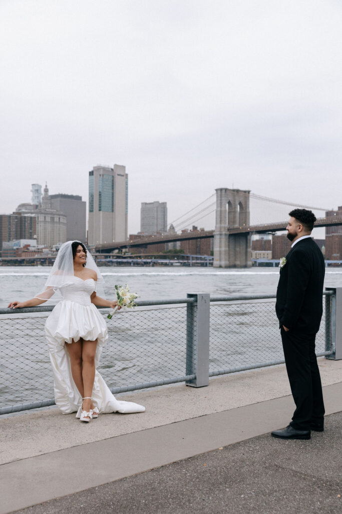 Bride leaning gracefully against a railing with the Brooklyn Bridge in the background, as the groom admires her from a distance