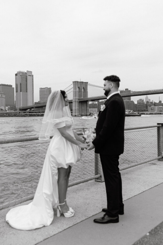 Black and white photo of a bride and groom holding hands by the Brooklyn waterfront, with the Brooklyn Bridge in the background