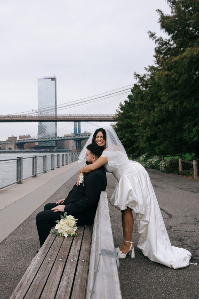 Bride joyfully hugging the groom from behind as he sits on a bench, with the Brooklyn Bridge and waterfront in the background