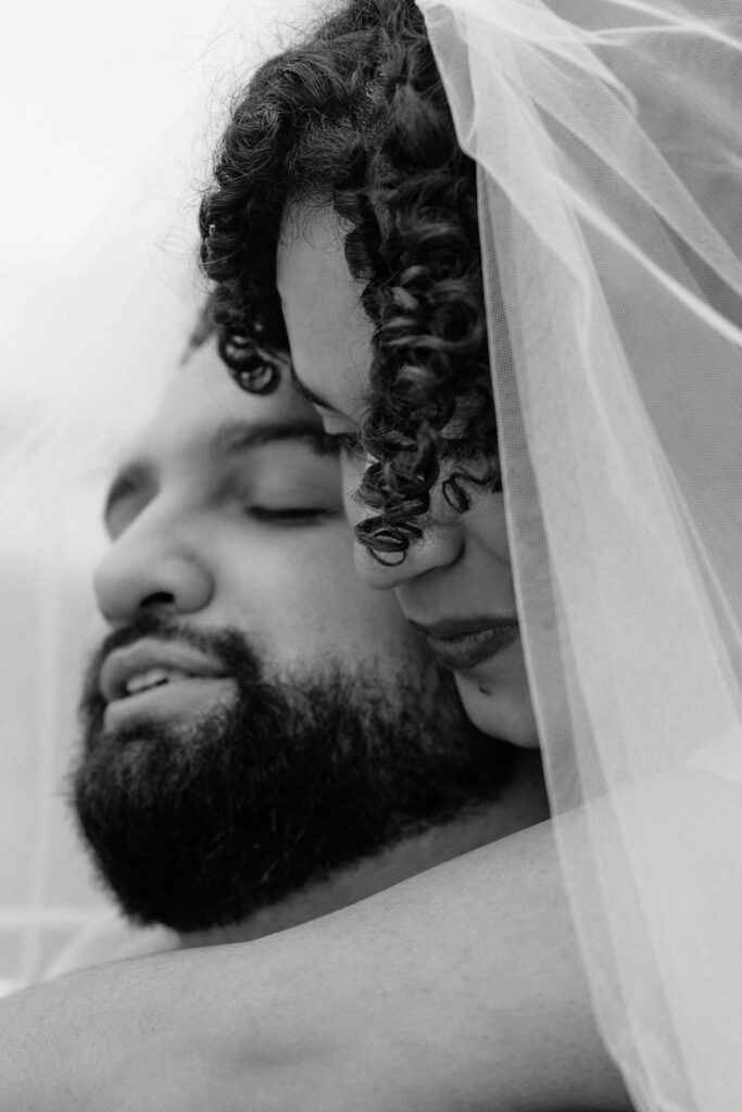 Black and white close-up of a bride and groom in a tender embrace, with the bride's veil softly framing their faces