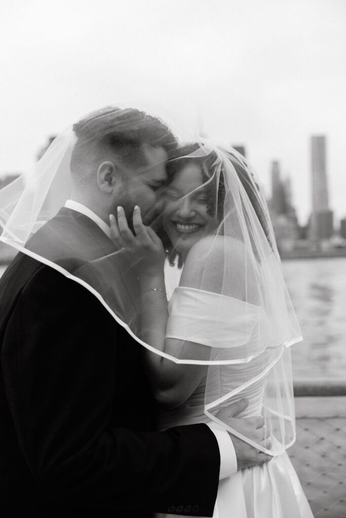 Black and white photo of a bride and groom sharing a joyful moment under the bride's veil, with the city skyline softly blurred in the background