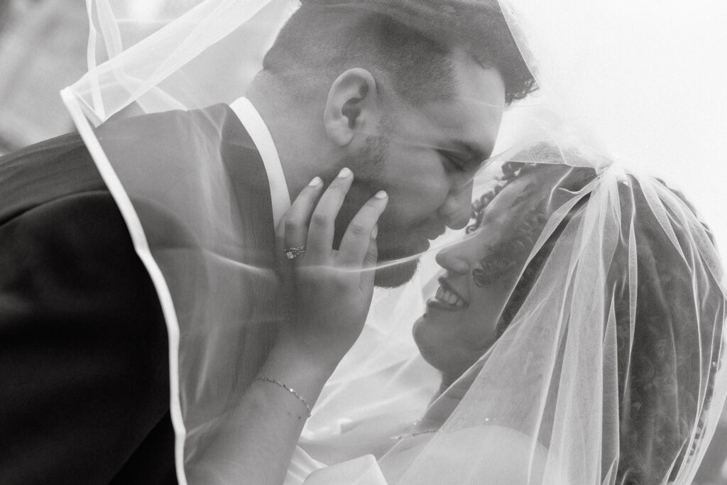 Intimate black and white close-up of a bride and groom smiling and leaning in for a kiss under the veil, highlighting the bride's engagement ring and their joyful expressions.