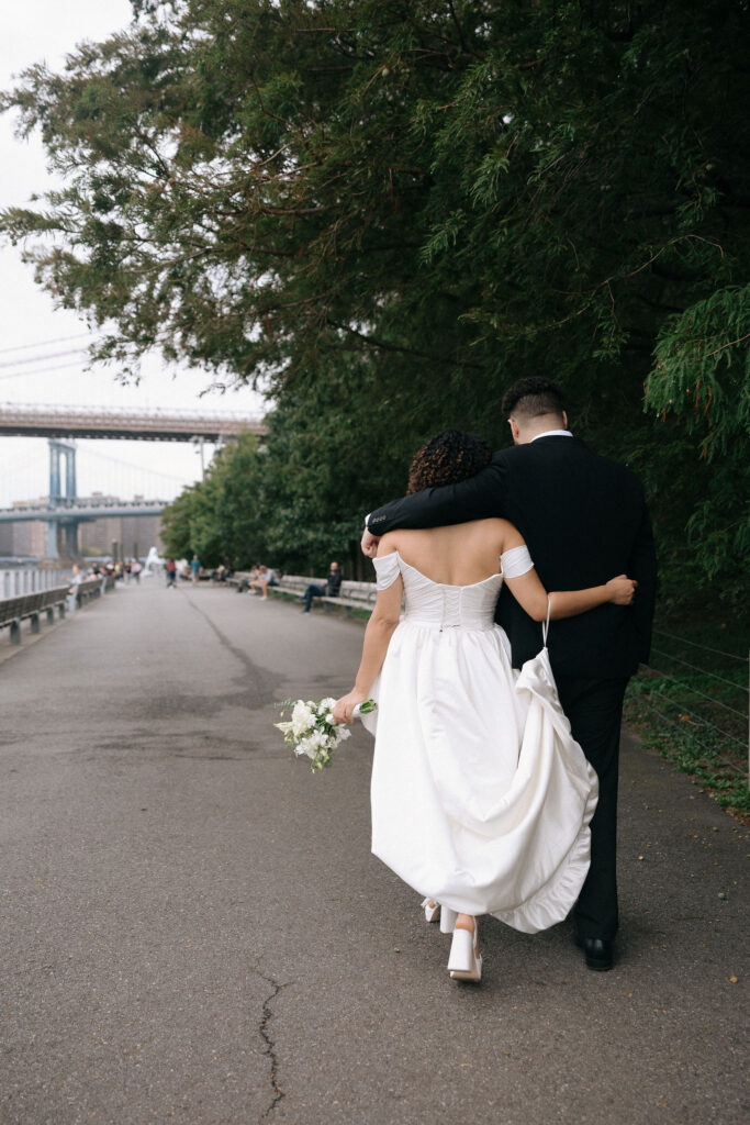  bride and groom walking hand in hand along a serene riverside path with the Manhattan Bridge in the background, the bride holding a bouquet and her gown flowing behind