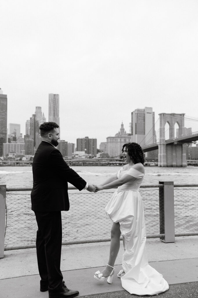 A black-and-white photo of a bride and groom holding hands and smiling at each other by the Brooklyn waterfront, with the city skyline and Brooklyn Bridge in the background