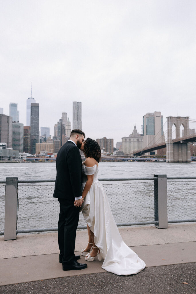 A bride and groom sharing an intimate moment by the Brooklyn waterfront, holding hands with the New York City skyline and Brooklyn Bridge in the background