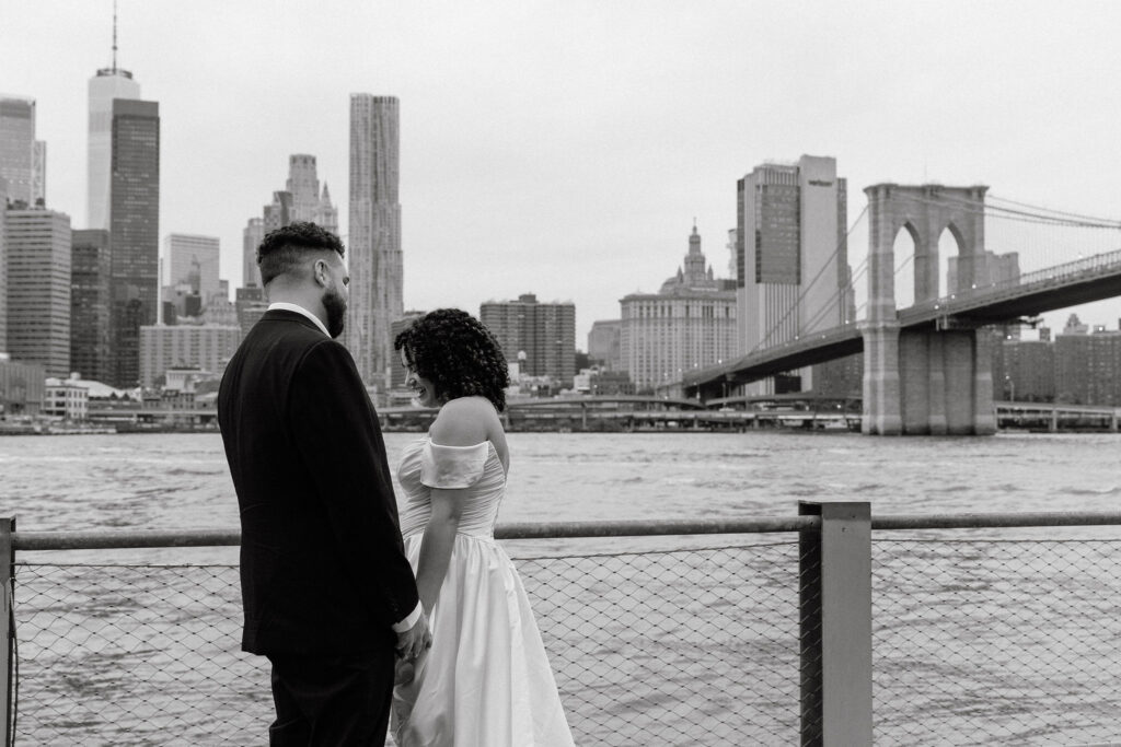 Couple standing by the waterfront with the Brooklyn Bridge and Manhattan skyline in the background, sharing an intimate moment.