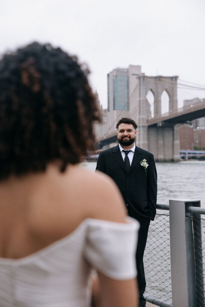 The groom, dressed in a black suit with a boutonniere, stands smiling in focus while the bride, blurred in the foreground, looks toward him near the Brooklyn Bridge