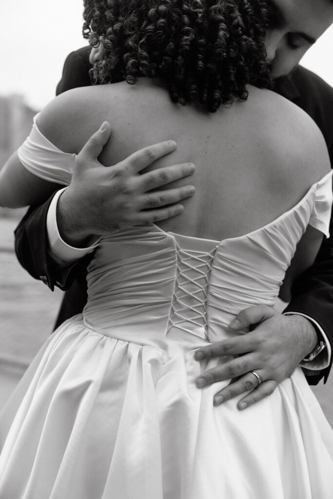 A close-up black-and-white shot of the groom tenderly embracing the bride, highlighting the intricate lace-up detail on her wedding dress and the groom's hand resting on her back