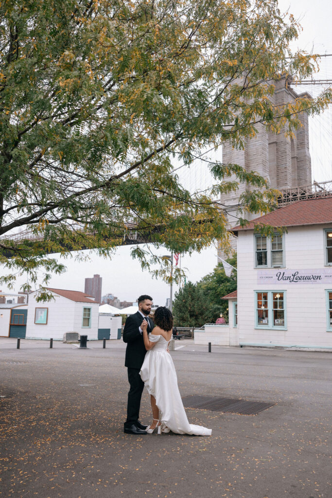 A bride and groom share a quiet dance under a large tree with golden leaves near the Brooklyn Bridge, with Van Leeuwen ice cream shop in the background.