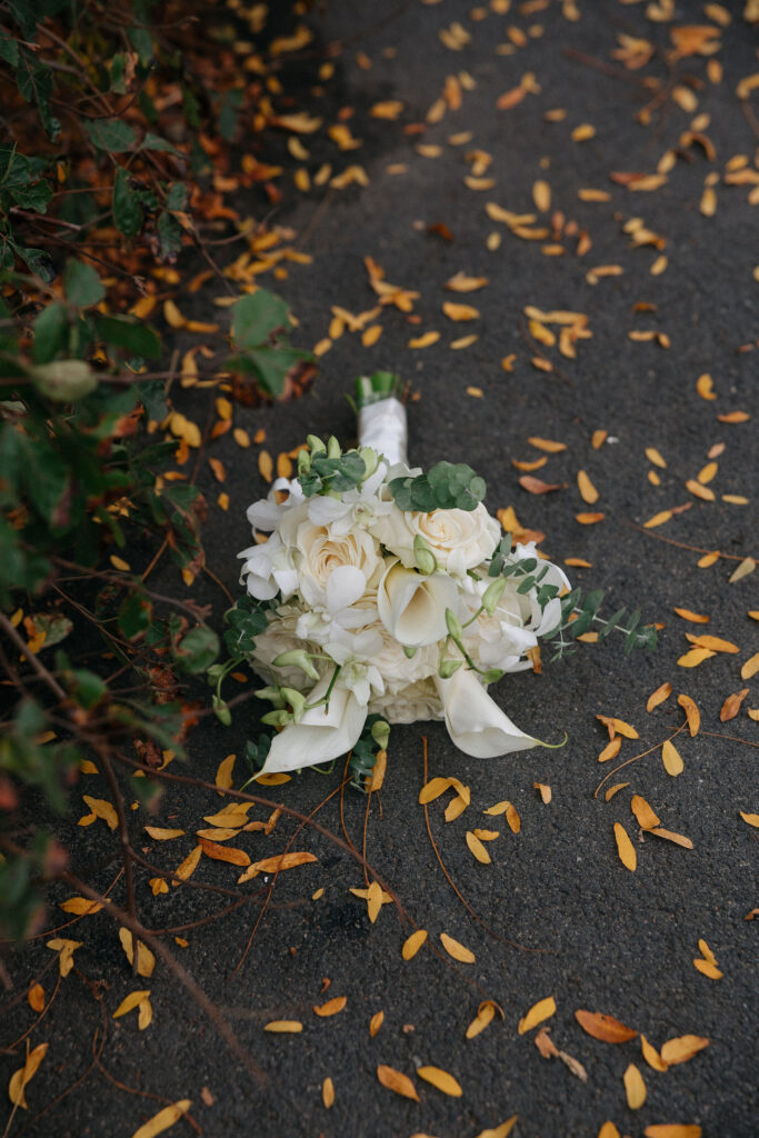 Close-up of a white bridal bouquet featuring roses, calla lilies, and eucalyptus leaves, resting on a path scattered with golden autumn leaves.