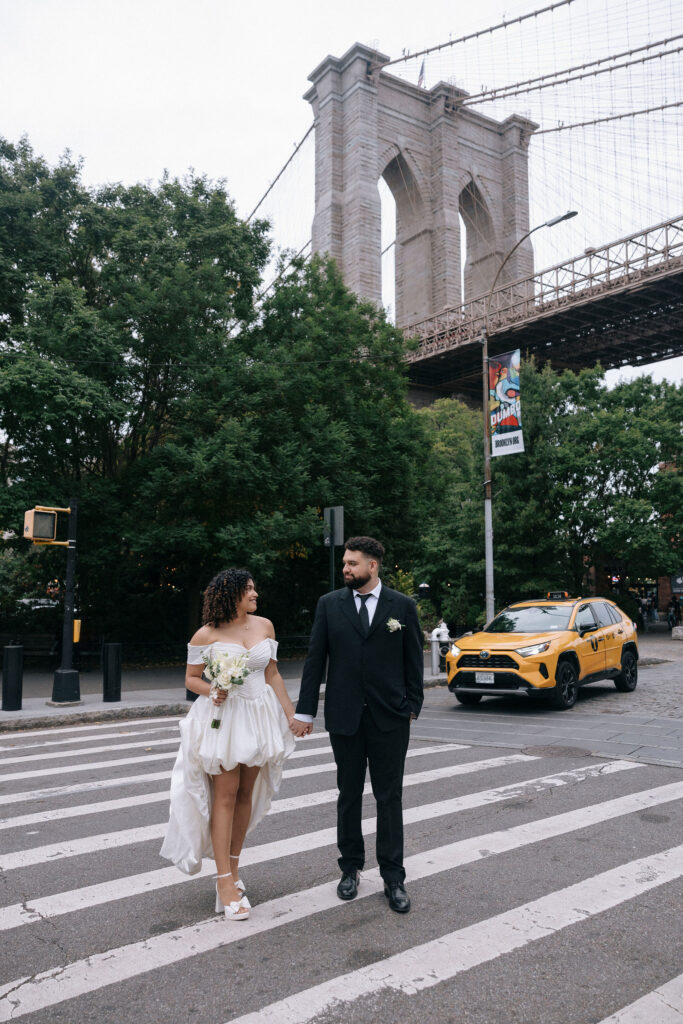 Bride and groom walking hand-in-hand across a crosswalk near the Brooklyn Bridge, with a yellow taxi in the background