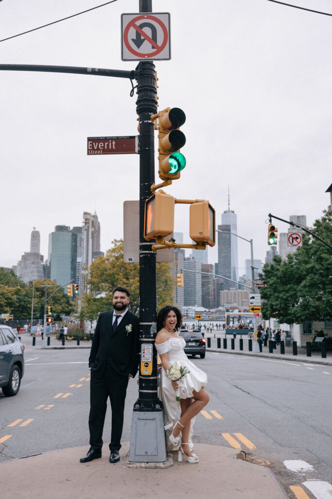 A bride and groom leaning against a traffic light pole at the corner of Everit Street with the New York City skyline in the background, capturing a fun and modern vibe.