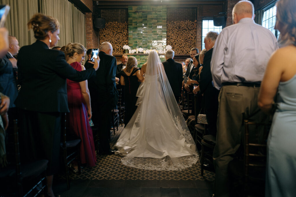 A bride walks down the aisle at the Bowery Hotel, her veil cascading behind her as guests look on in admiration during an intimate wedding ceremony.