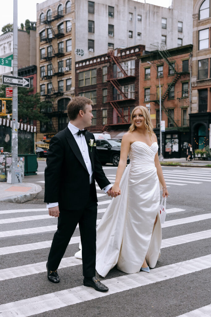 Text: A newlywed couple walks hand-in-hand across a New York City crosswalk, dressed in classic wedding attire. The groom wears a black tuxedo, and the bride glows in a satin gown, smiling at him as they stroll through the city streets.