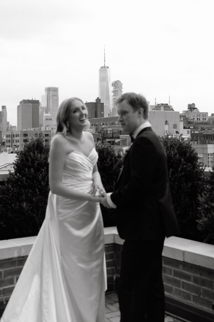 A joyful bride and groom share a candid moment on the rooftop of the Bowery Hotel, holding hands with the Manhattan skyline in the background.