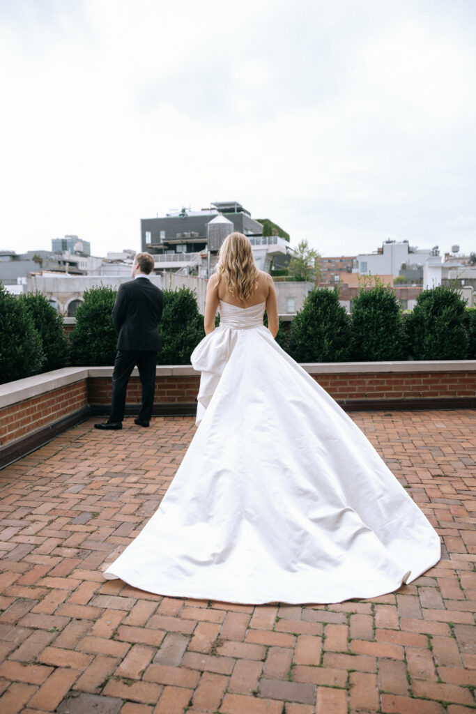 A bride in a stunning white gown walks towards her groom for their first look on the rooftop of the Bowery Hotel, with the NYC skyline in the background.