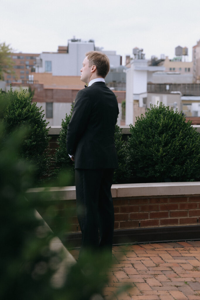 A groom stands on the rooftop of the Bowery Hotel, waiting in quiet anticipation for his bride during their first look, with the NYC skyline behind him.