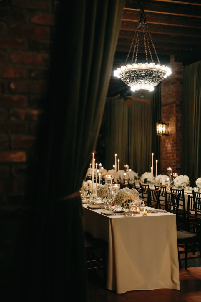 An elegant wedding reception setup at the Bowery Hotel, featuring candlelit tables, white floral arrangements, and a vintage chandelier casting a warm glow.