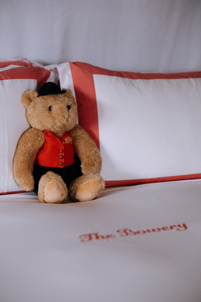 A signature Bowery Hotel teddy bear sits on a neatly made bed with embroidered sheets, adding a charming and cozy touch to the wedding day preparations.