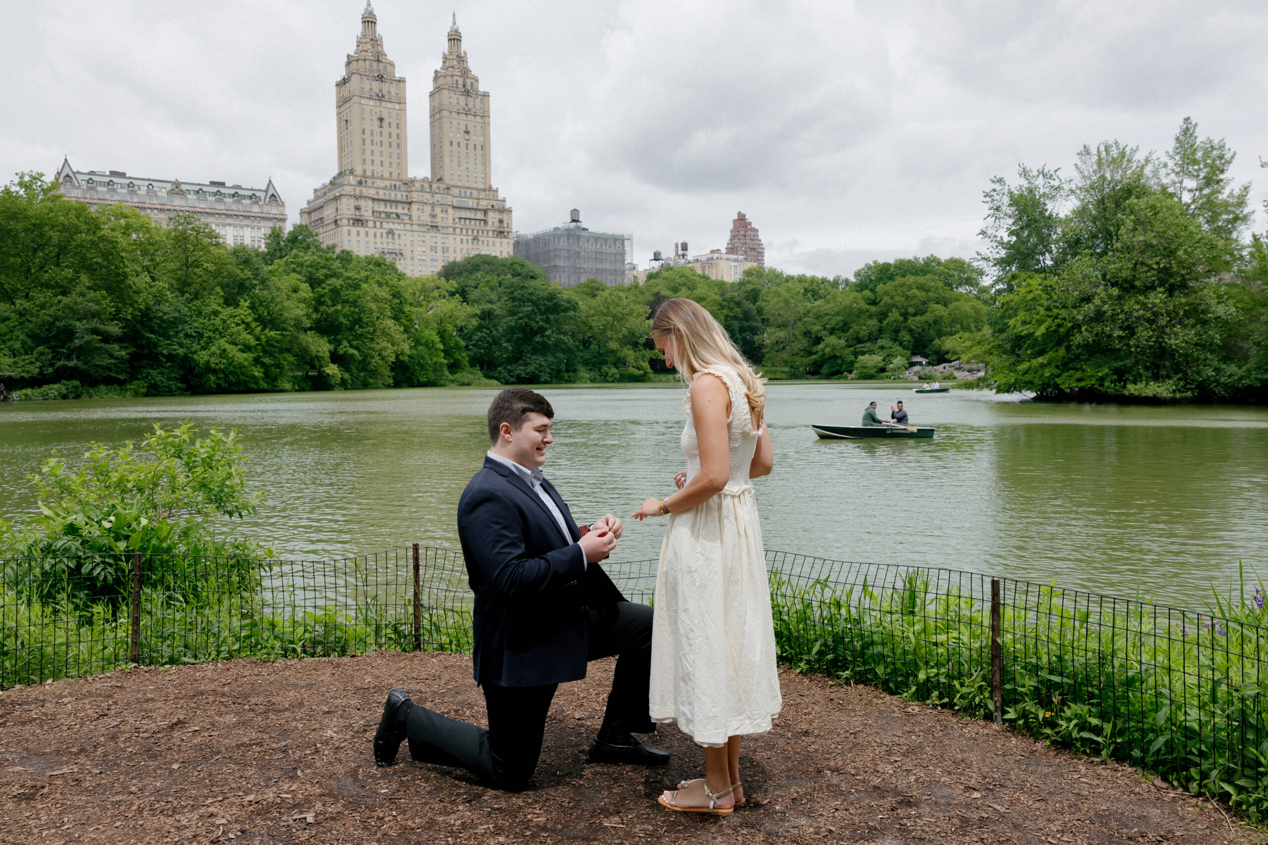 Surprise proposal in Central Park by the lake with Bow Bridge and San Remo building in background, NYC engagement photographer captures candid moment