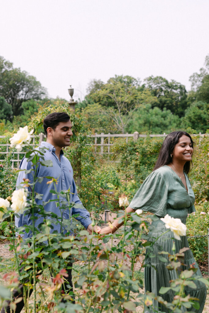Candid engagement photo of a couple laughing in the Rose Garden at Brooklyn Botanic Garden, captured by NYC proposal photographer.
