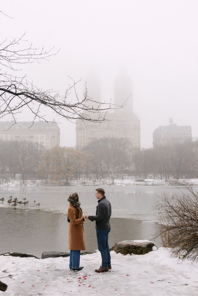 couple standing by a snowy lake in foggy central park with the manhattan skyline in the background