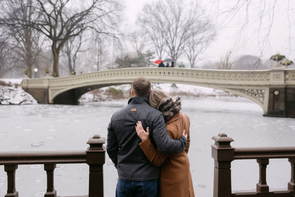 engaged couple hugging in front of a snowy bridge in central park after a surprise proposal