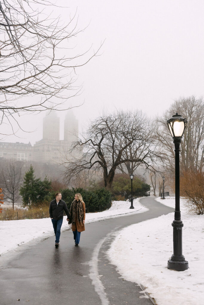 engaged couple walking away together on a foggy winter path in central park after a surprise proposal