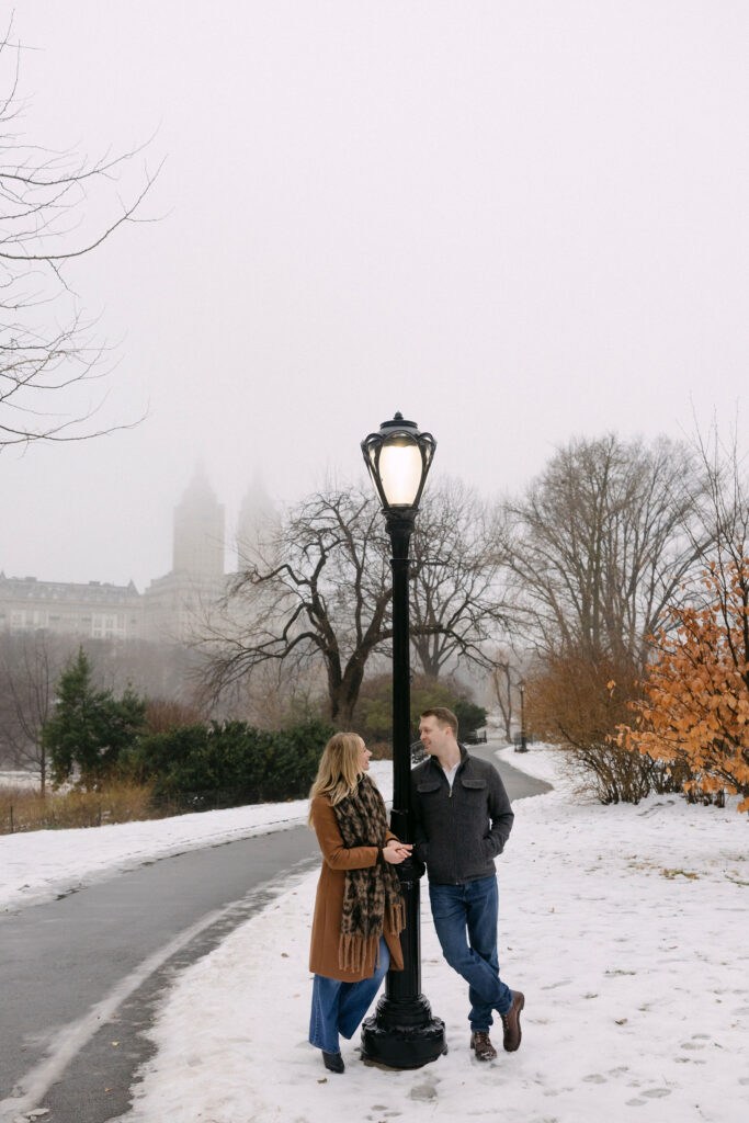 engaged couple standing together on a snowy path in central park near a lamppost on a foggy winter morning