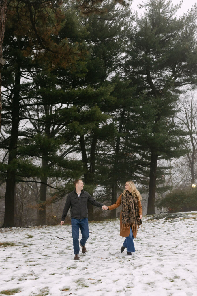engaged couple walking and laughing together through snowy trees in central park after a surprise proposal