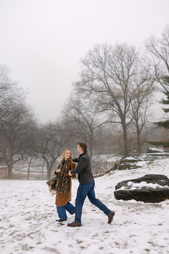 engaged couple walking together across a snowy open area of central park after a surprise proposal