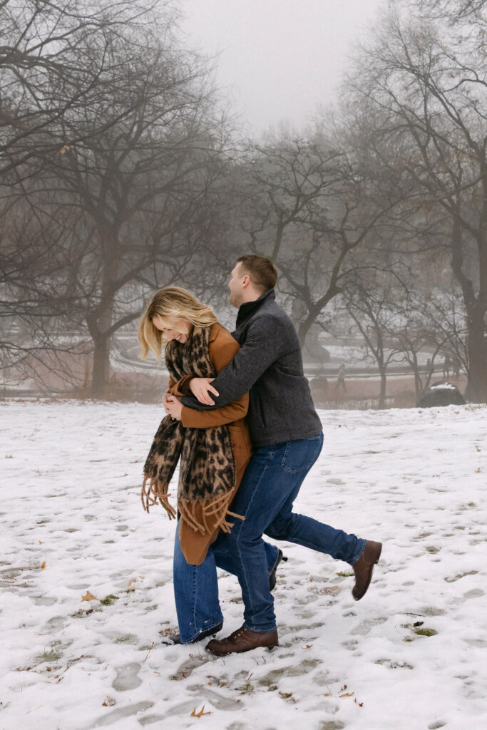 engaged couple laughing and hugging in the snow in central park after a surprise proposal