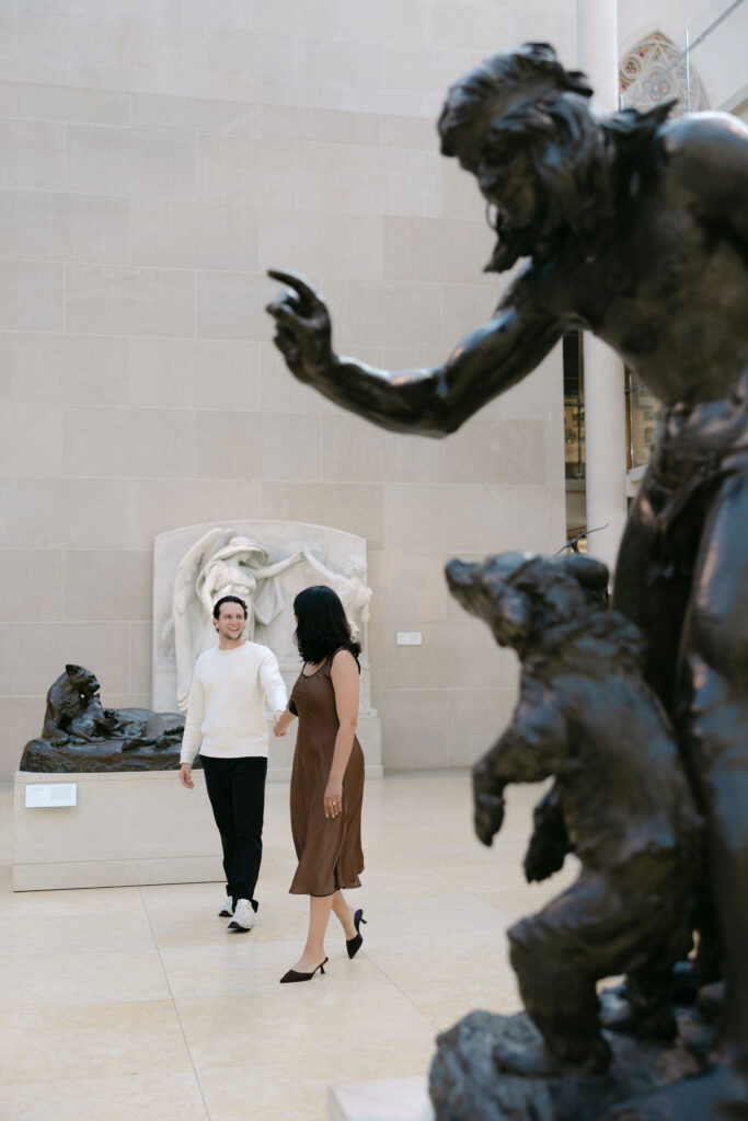 couple walking past a sculpture inside a museum near central park during an nyc engagement session