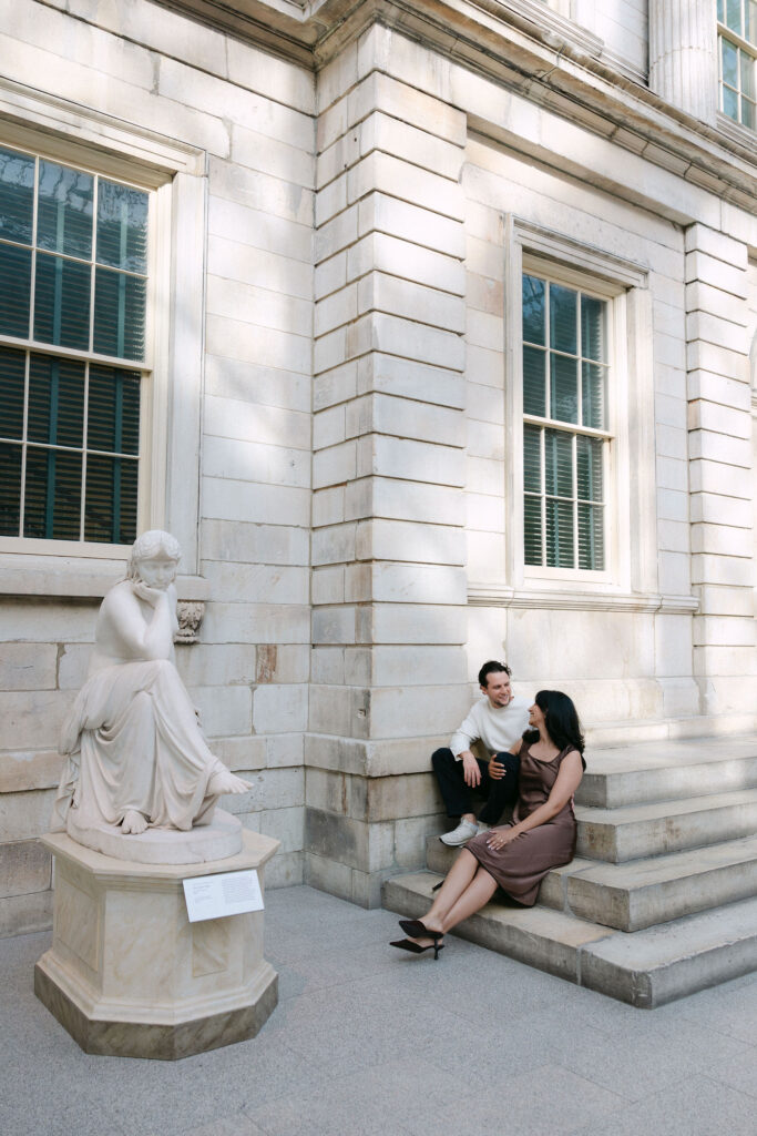 nyc engagement session photo of a couple sitting on steps near central park surrounded by classic new york architecture
