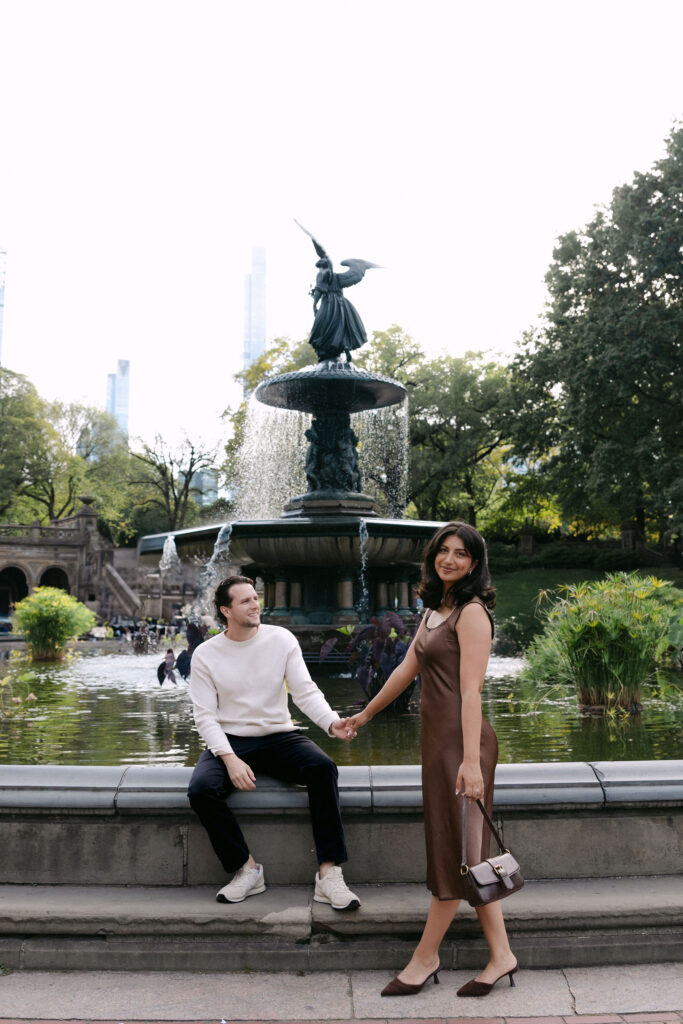 nyc engagement session photo of a couple sitting and standing near a fountain in central park